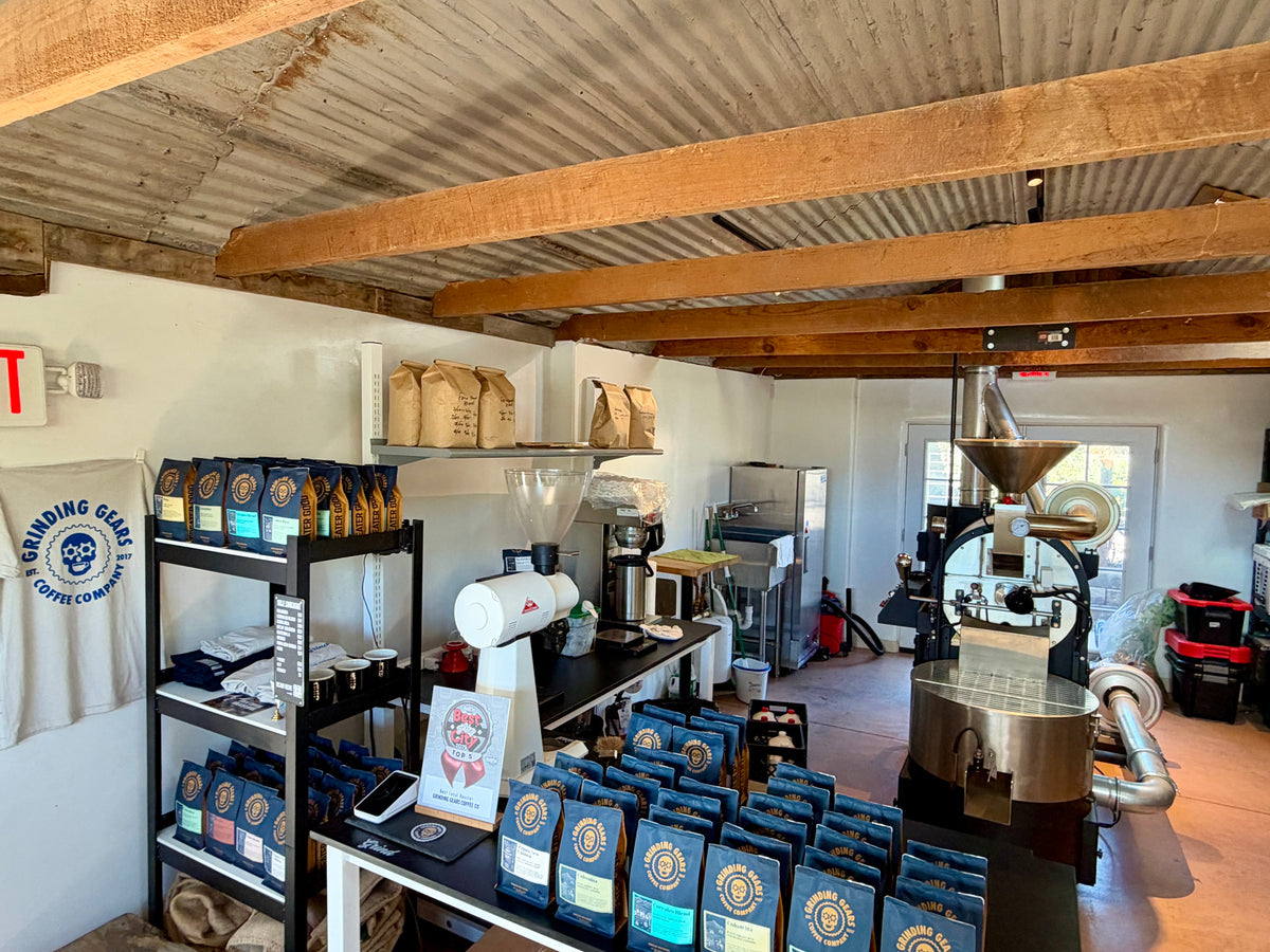 Inside the Grinding Gears Coffee roastery at the Farm Stand in Corrales, with the coffee roaster, grinder, and bags of freshly roasted coffee beneath wooden beams.